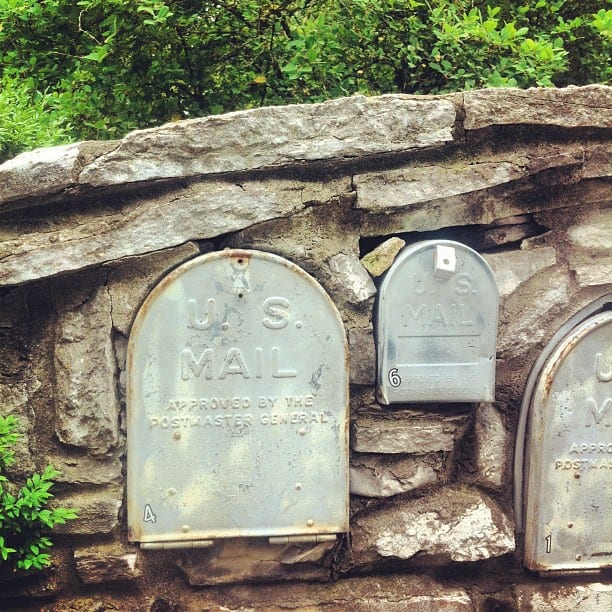 Vintage silver U.S. Mail mailboxes nestled into a rustic stone wall surrounded by green foliage.
