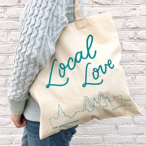 Model wearing market bag over shoulder. Bag shows handlettered words Local Love and a line drawing of the skyline of Charleston, SC