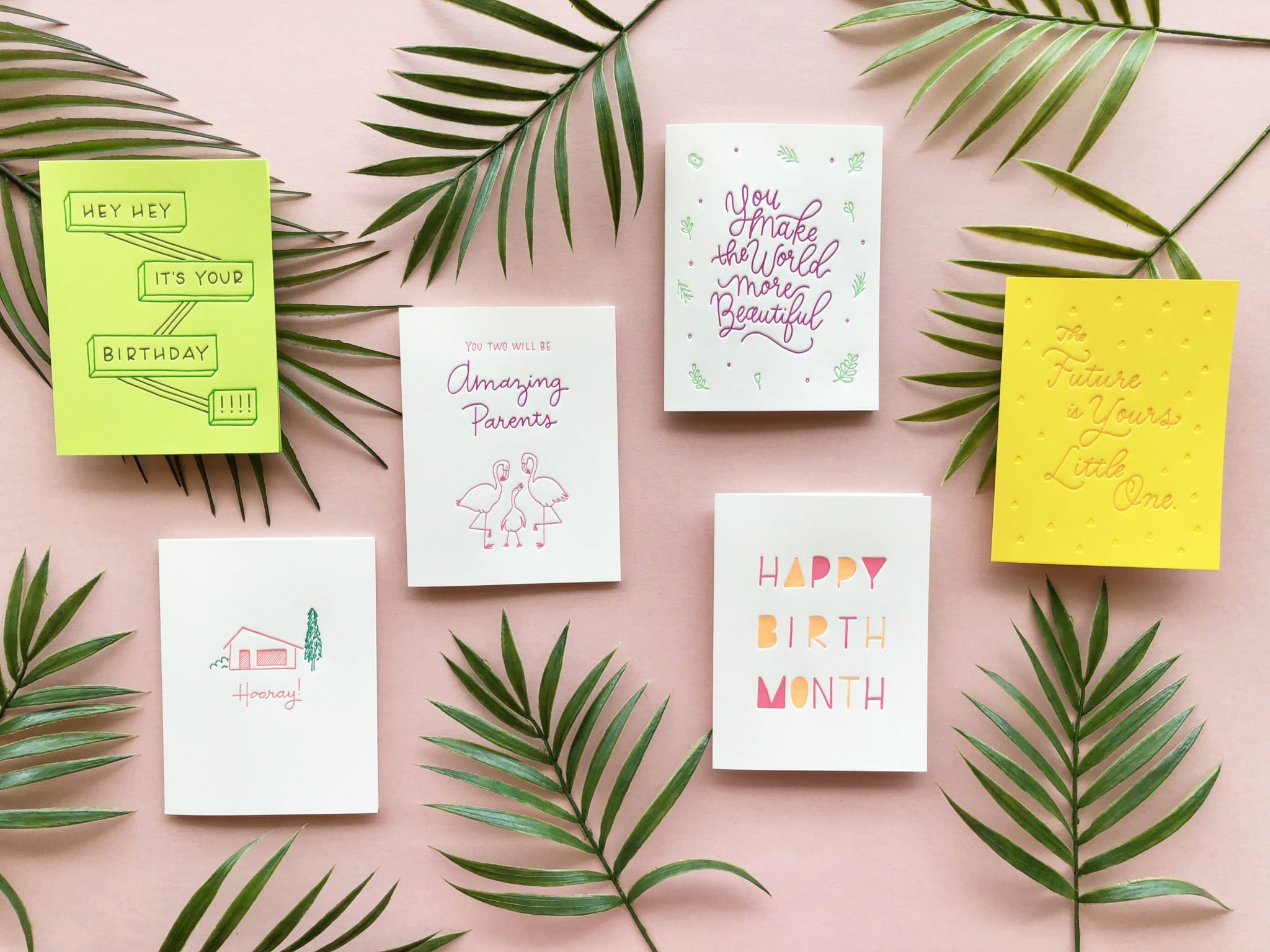 Colorful greeting cards arranged on a light pink background with palm leaves