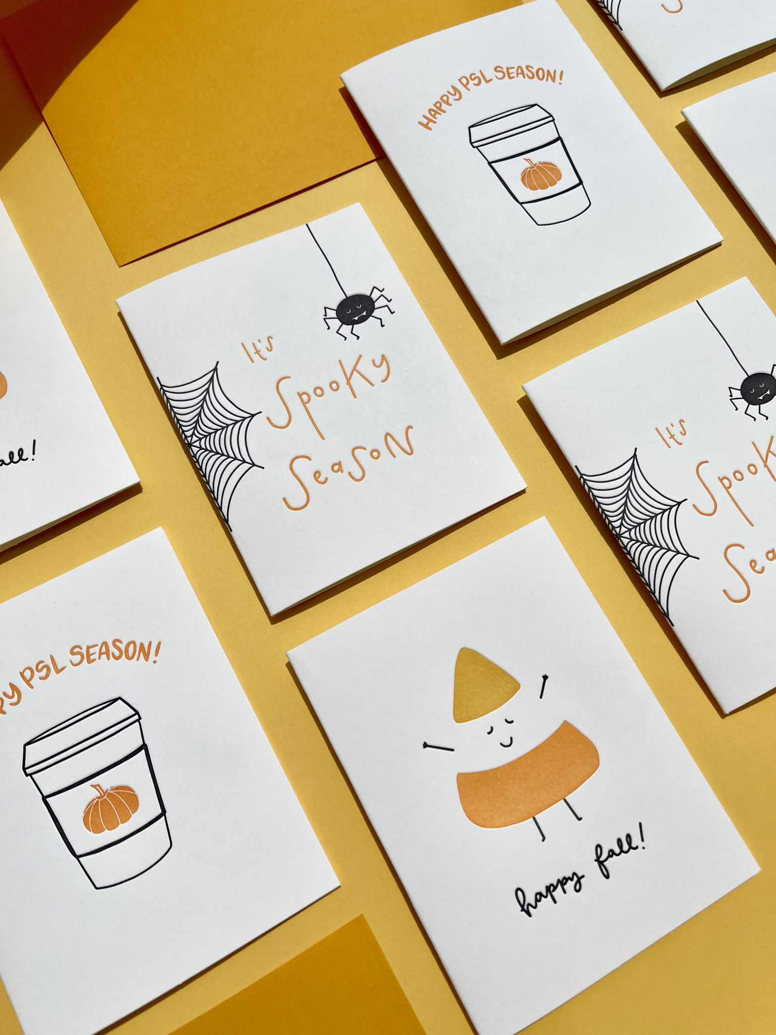 Assorted Halloween and fall greeting cards featuring spiders, candy corn, and pumpkin spice latte designs on a yellow background.